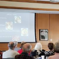 A man speaks to a crowd with a presentation displaying coins